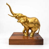 Goldfarbene Bronzefigur eines Elefanten mit erhobenem Rüssel und Stoßzähnen auf einem rechteckigen braunen Sockel.