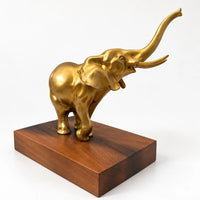 Schräge Rückansicht der goldfarbenen Elefantenfigur mit erhobenem Rüssel auf braunem Sockel.