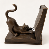 Katze aus Bronze mit Brille und hochgeschwungenem Schwanz vor einem aufgeschlagenen Buch, in Seitenansicht.